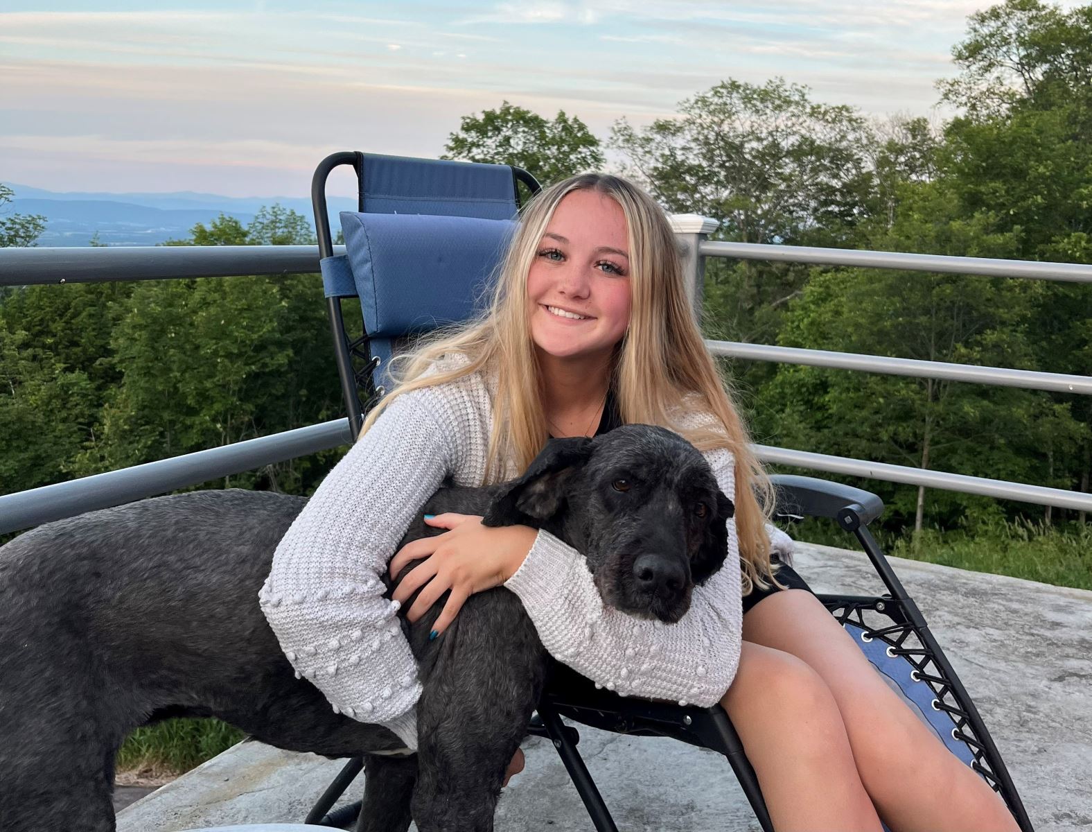 High school graduate sits on a deck with her brown dog overlooking the mountains